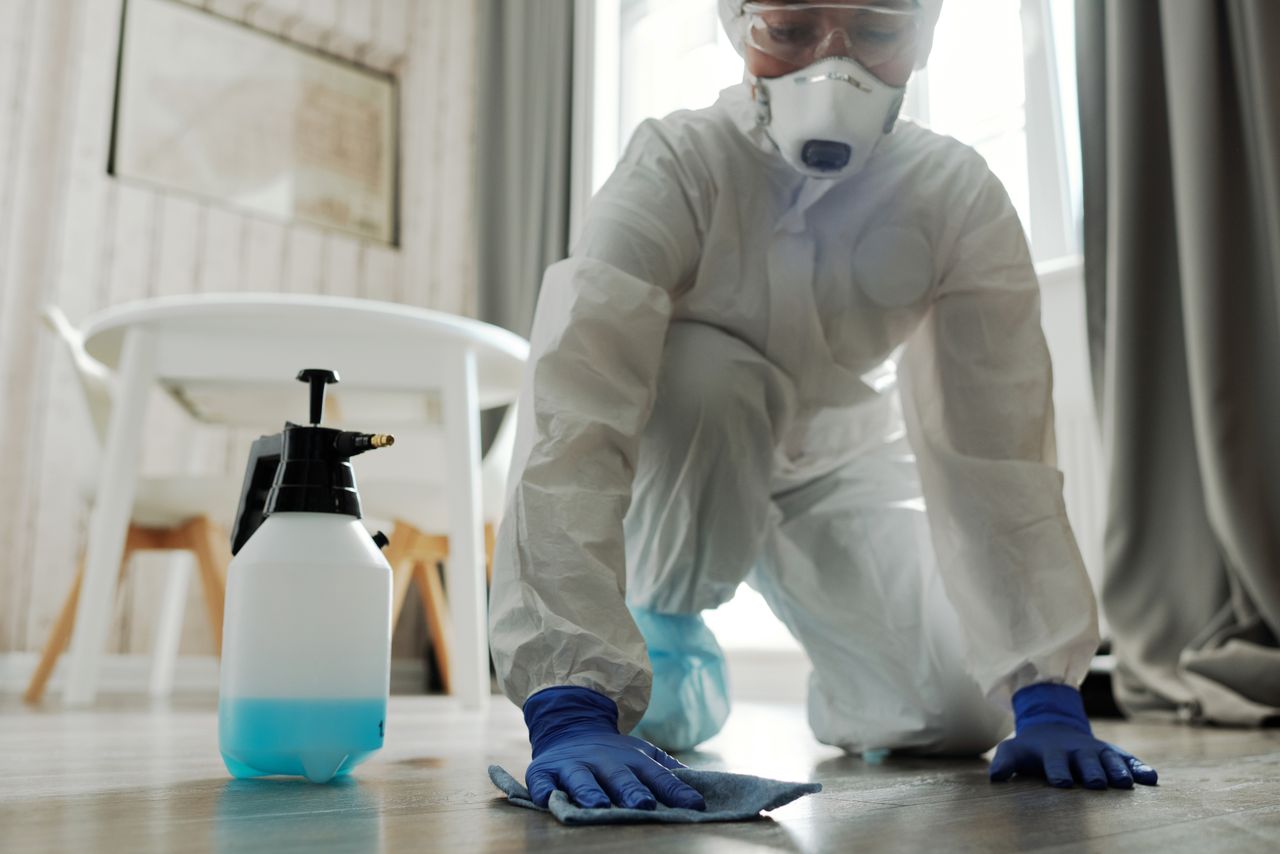 Person in protective gear cleaning a floor with disinfectant spray and cloth indoors.
